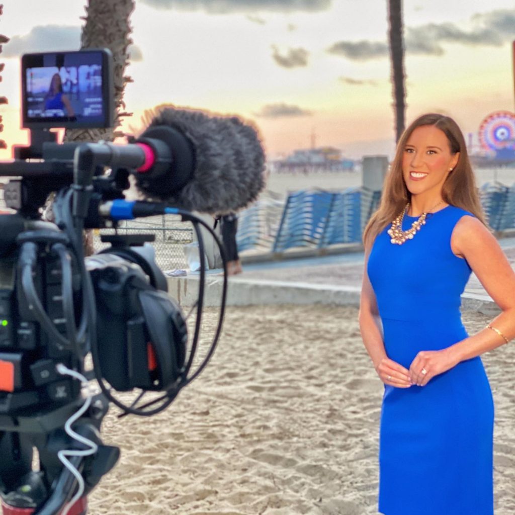 Filming in Santa Monica - Maggie Clark wears a blue dress on the Santa Monica Beach as a camera operator prepares to film her with the Santa Monica Pier | Photo by Magie Clark, @MaggieClarkOnCamera