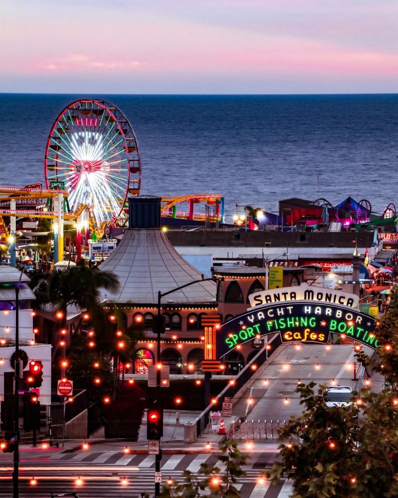 An image of the Santa Monica Pier at dusk as seen from Downtown Santa Monica at dusk with a snow man depicted in lights on the Ferris wheel. Photo by @kauaidreamerimages